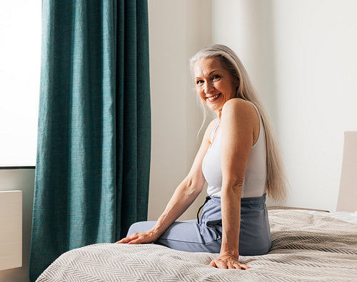 Smiling senior woman with long white hair looking at the camera while sitting on a bed. Female in casuals in bedroom.