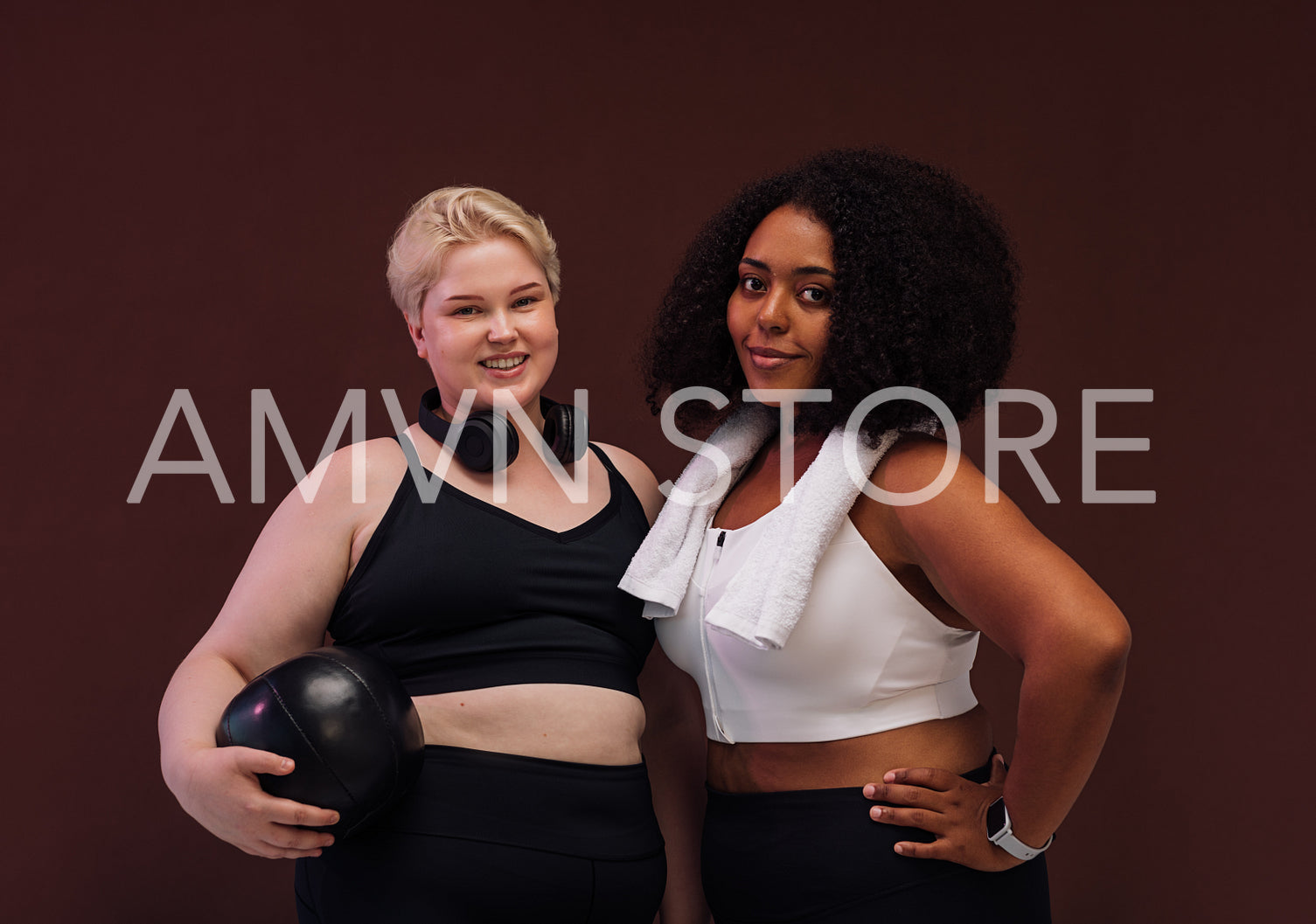 Two cheerful women in sportswear posing on brown background. Plus size females standing together with fitness equipment.