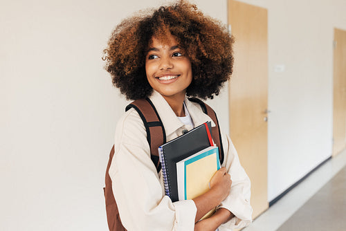 Smiling student with curly hair standing in corridor and looking away. Girl with textbooks and laptop in school.