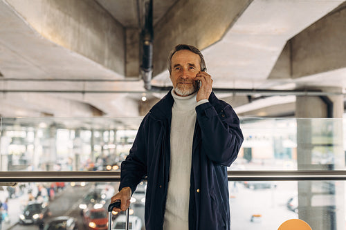 Senior tourist standing in airport terminal with luggage and talking on cell phone