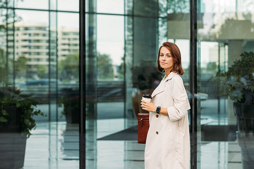 Stylish businesswoman standing at glass building holding a coffee and folder