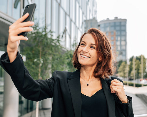 Smiling middle-aged businesswoman with ginger hair taking a selfie outdoors on her smartphone