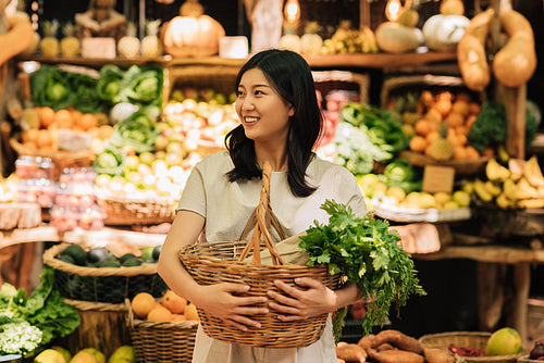 Smiling Asian woman holding wicker shopping basket with vegetables while standing at the farmers market