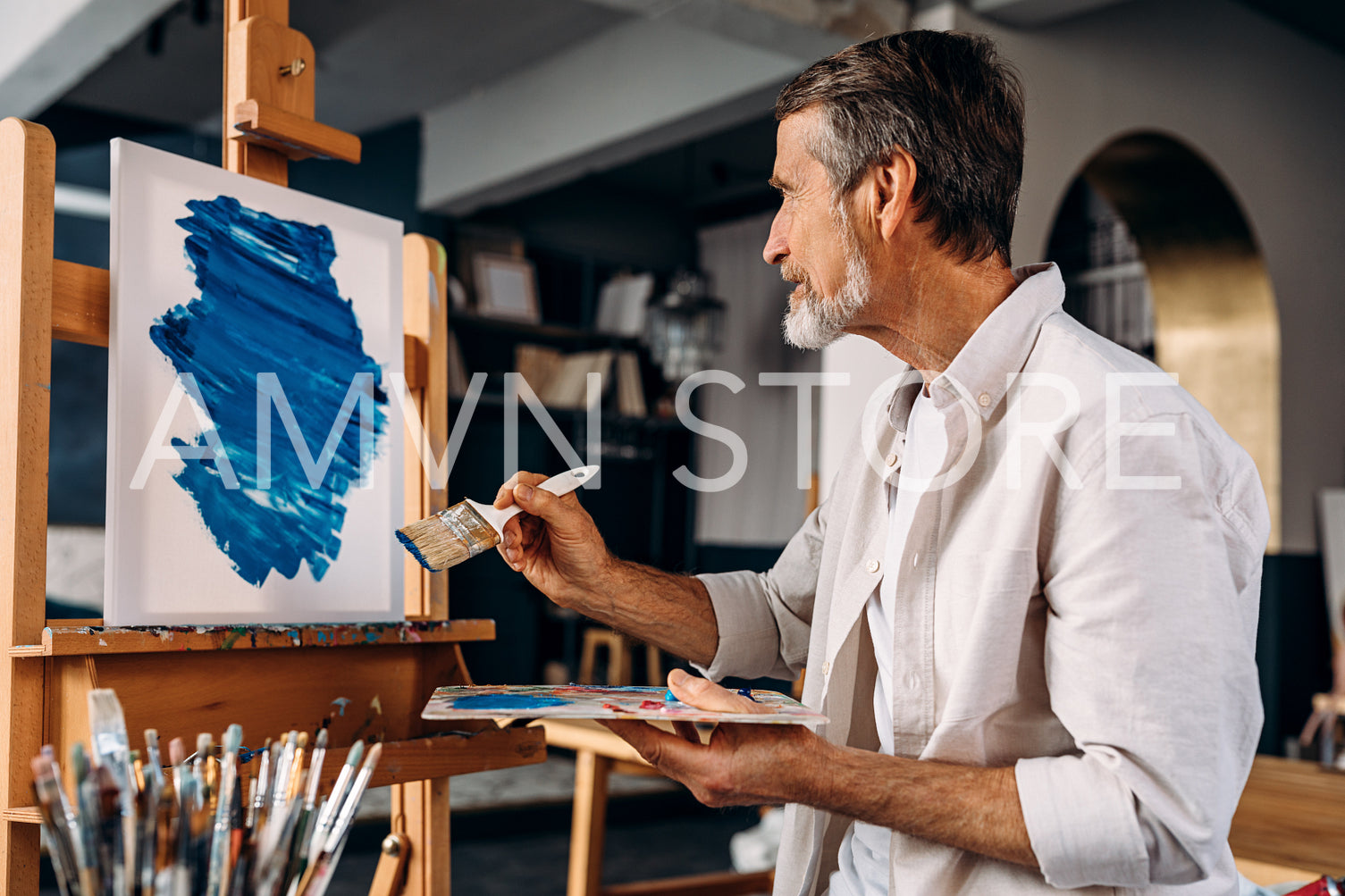 Bearded artist sitting in front of easel and painting abstract picture with large paintbrush in studio	