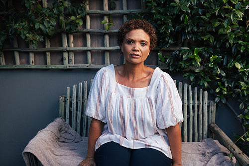 Portrait of a mature woman with short hair looking at camera while sitting on a bench at backyard of her house