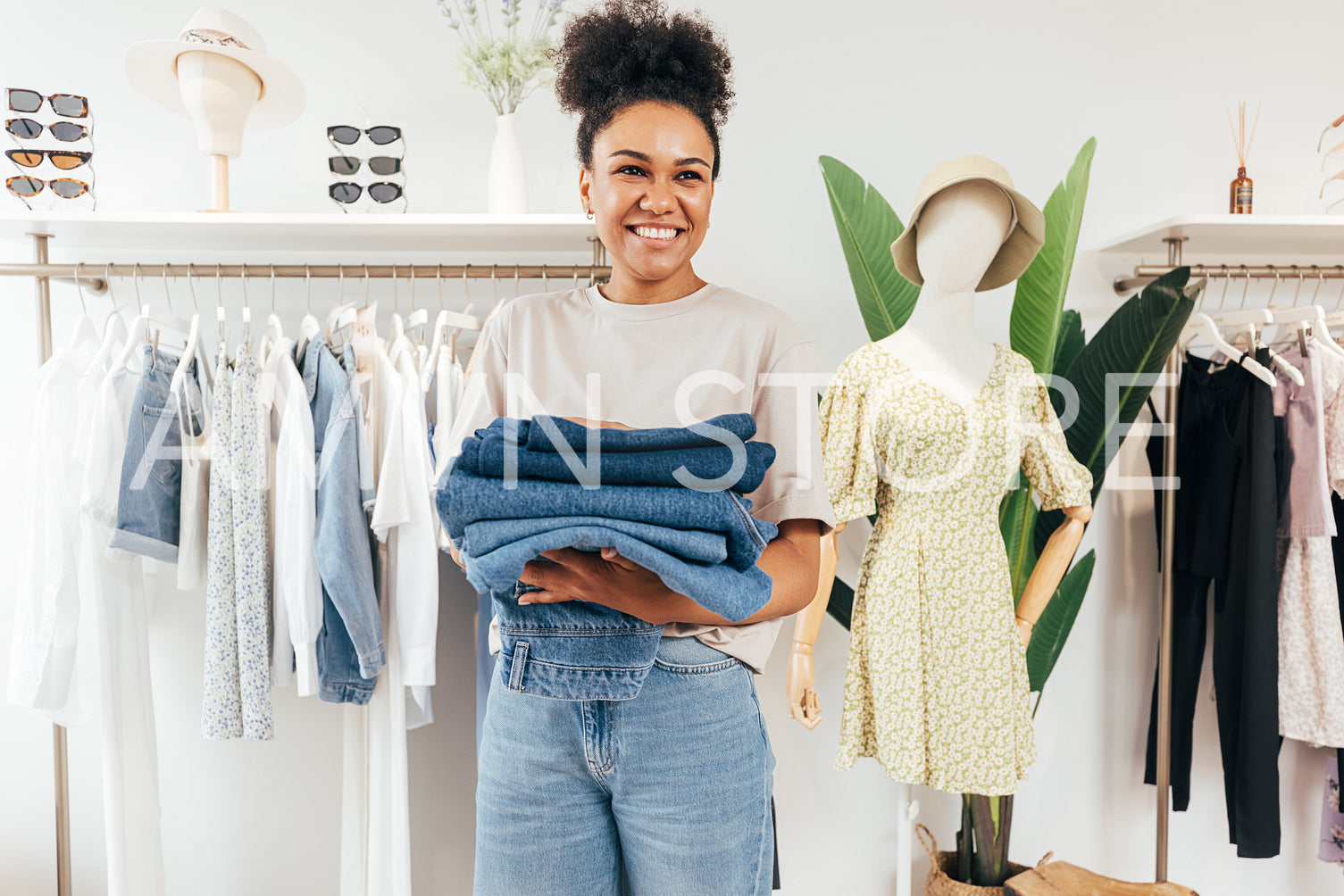 Portrait of a smiling woman in her small clothing retail shop. Happy business owner holding a pile of clothing and looking away.