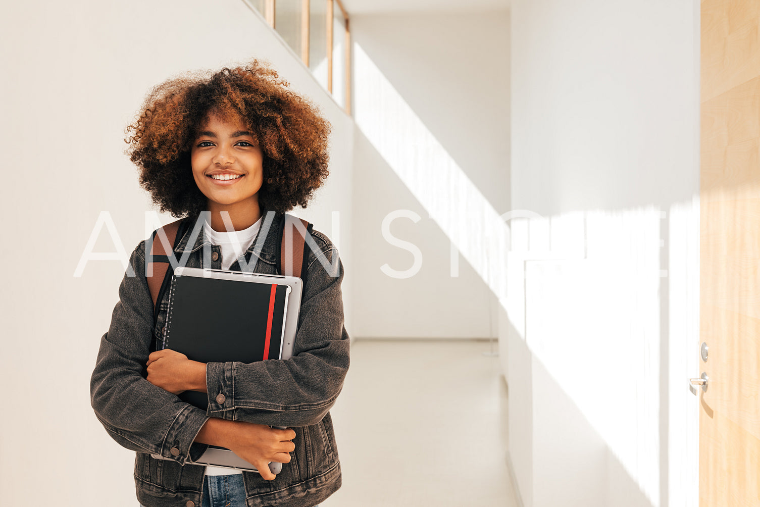 Portrait of a smiling student holding notebook and laptop while standing in corridor