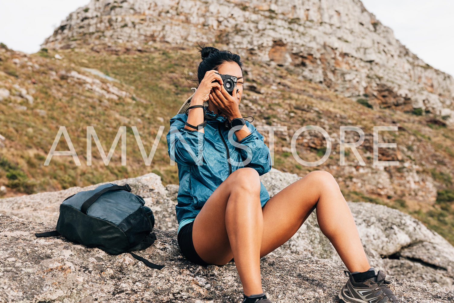 Fit woman in sports clothes with backpack sitting on stone taking photographs on an analog camera