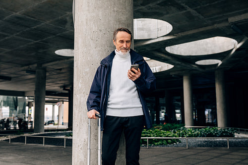 Senior man with rolling suitcase standing outdoors looking at mobile phone