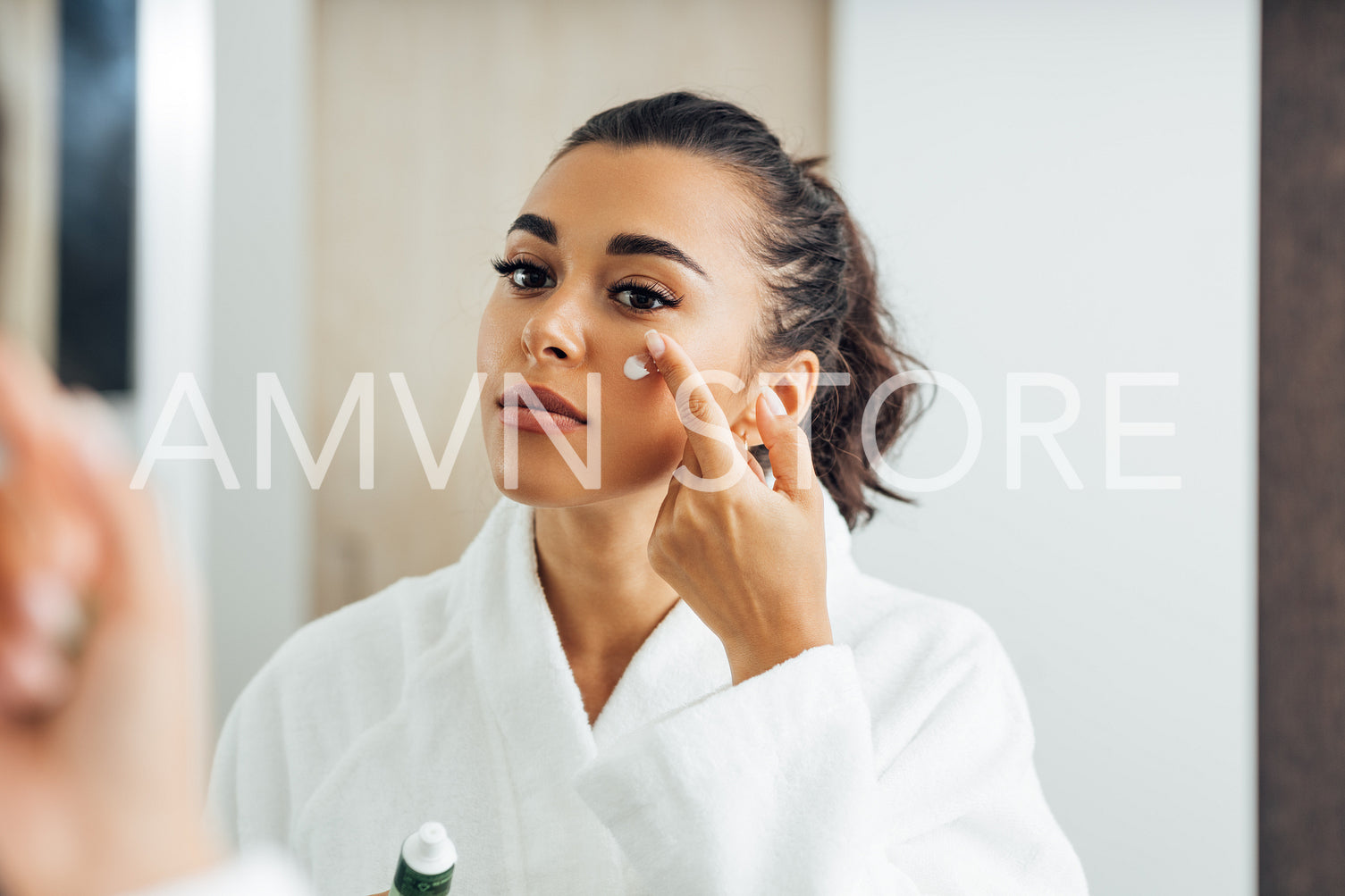 Beautiful woman in bathrobe applying anti-aging cream on her cheek	