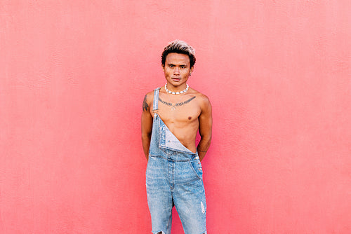 Portrait of a young handsome guy wearing jeans overalls standing against pink wall