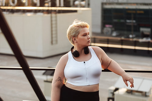 Woman with oversized body leaning to a railing during a workout on a roof. Oversize fitness woman in sportswear wearing headphones looking away outdoors.