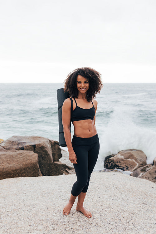 Beautiful smiling woman standing with yoga mat by ocean and looking at camera