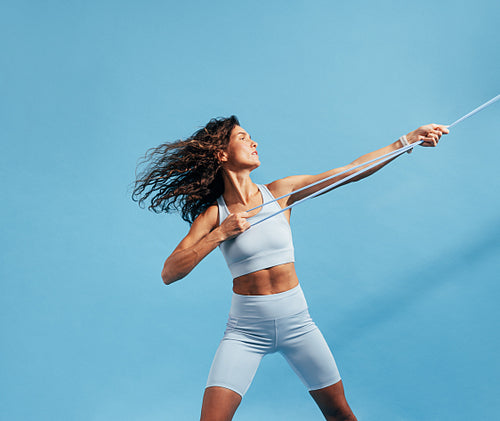 Woman in sportswear doing intense training with a resistance band. Slim female in sportswear exercising on blue background.