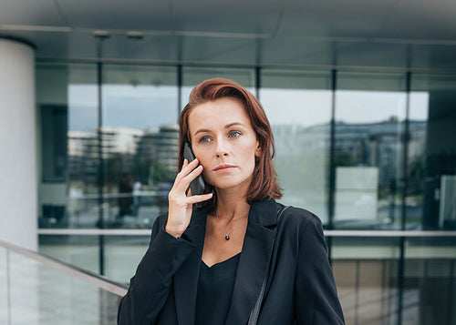 Portrait of a middle-aged female with ginger hair talking on her mobile phone while standing outdoors against a glass building