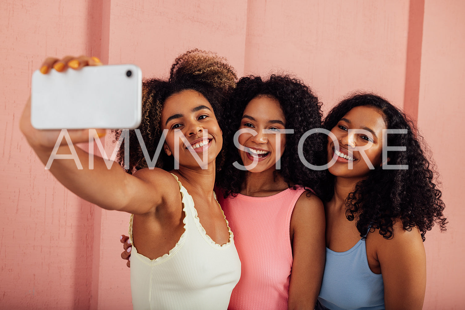 Young women in bright casuals standing together and taking selfie on a mobile phone. Happy females having fun against pink wall outdoors.