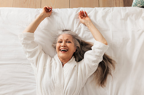 Happy senior woman in a bathrobe lying on a bed in a hotel room looking at camera