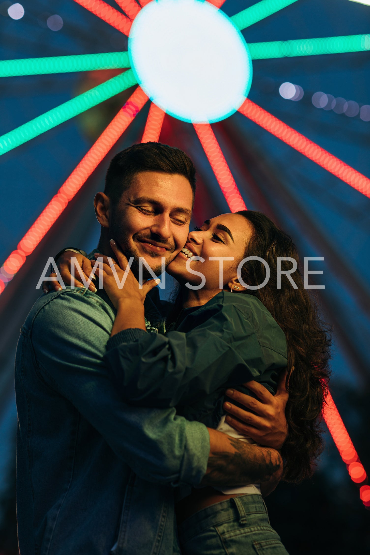 Young happy couple against Ferris wheel during the festival. Woman kissing her smiling boyfriend at night.