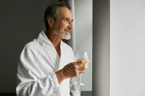 Smiling senior man with wineglass. Side view of a bearded mature male wearing a bathrobe in a hotel room looking at window.