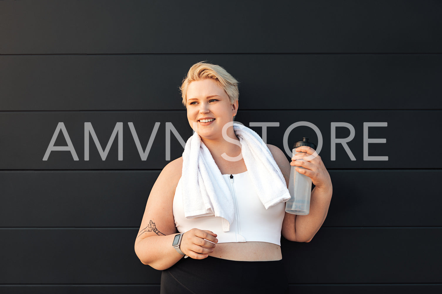 Smiling plus size woman sportswear standing at black wall. Healthy woman relaxing after training holding a bottle of water looking away.	