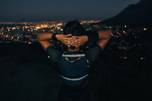 Woman relaxing after workout and looking at the cityscape at night. Rear view of athletic female standing with his hands behind head and taking a break.