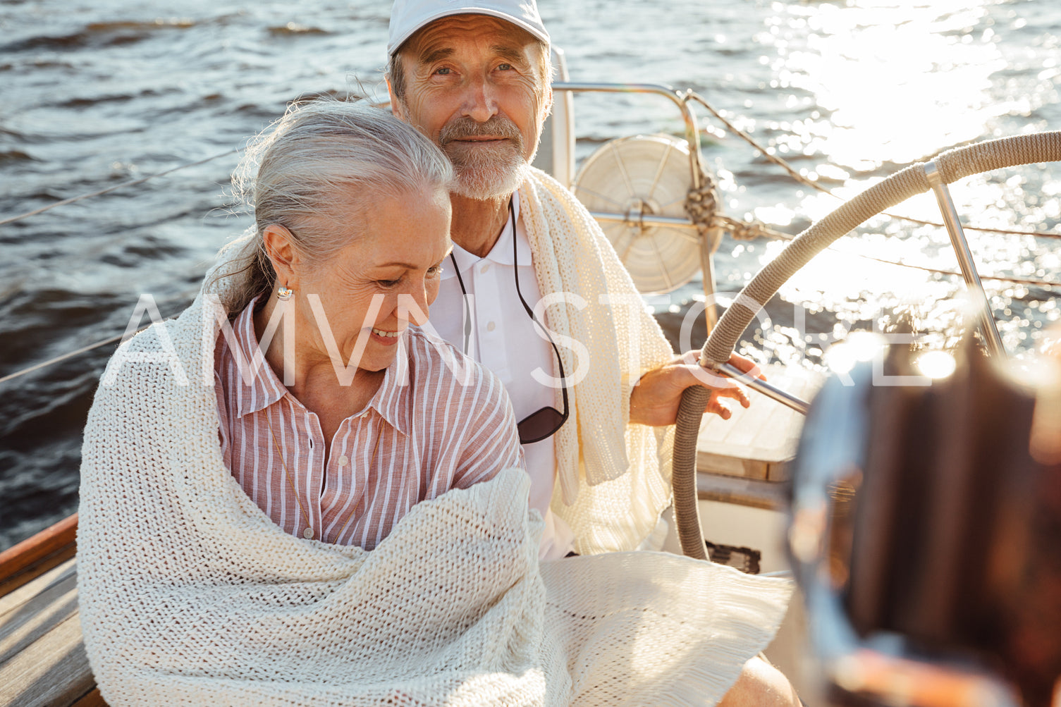 A couple of two mature people wrapped in plaid on a sailboat. Senior man in cap steering a yacht.	