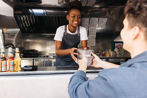 Young woman in apron working at food truck. Small business owner giving takeaway packaged food to customer.