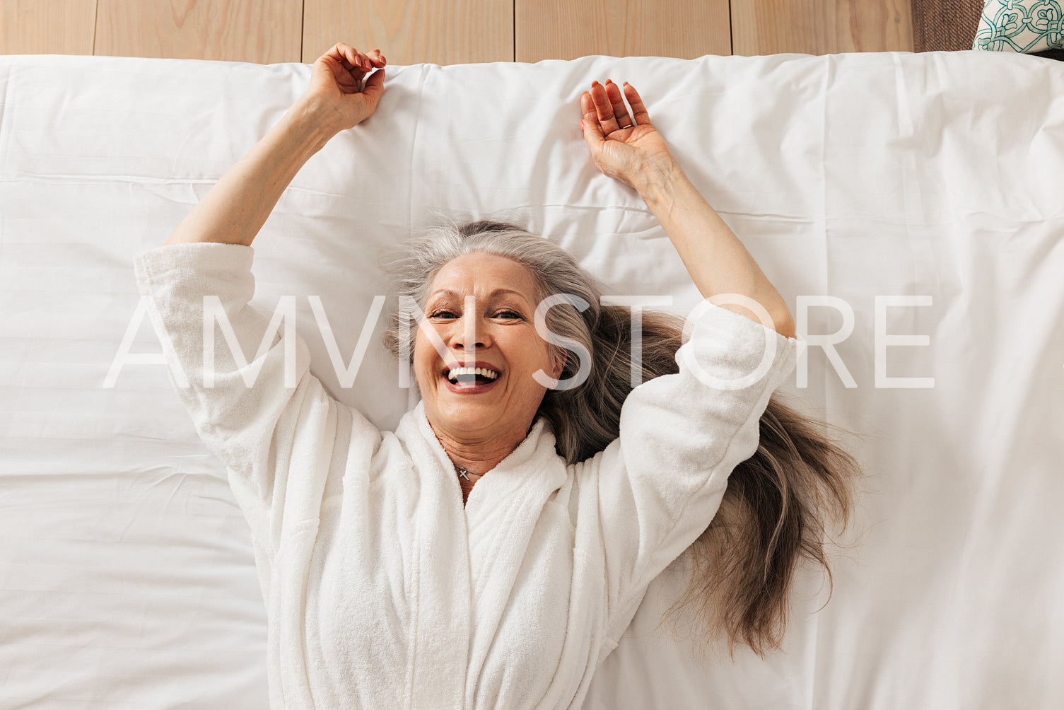 Happy senior woman in a bathrobe lying on a bed in a hotel room looking at camera