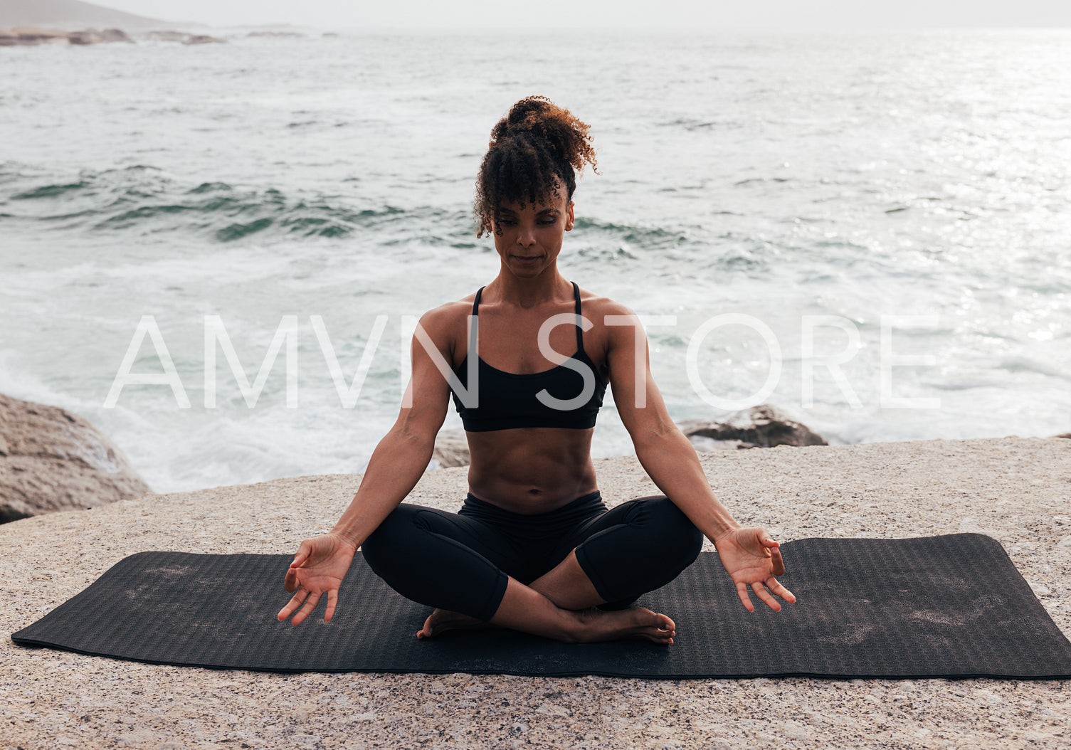 Woman in lotus pose sitting on yoga mat by the ocean at sunset