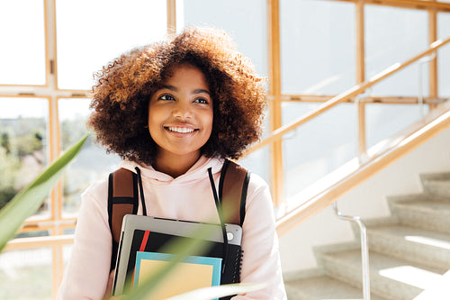 Cheerful girl with backpack holding textbooks. Young female smiling and looking away in school.