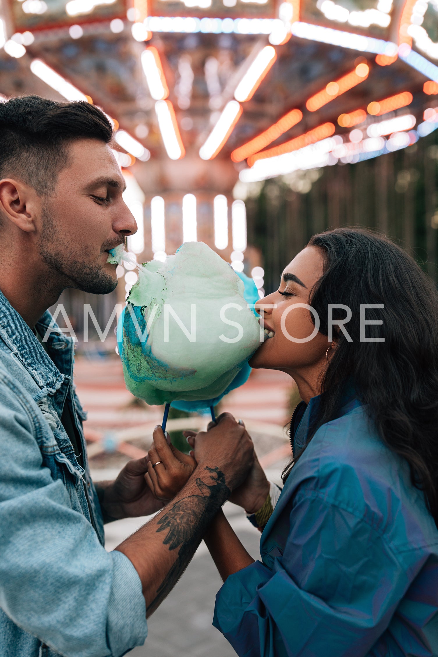 Side view of a young couple eating cotton candy against a carousel during the festival