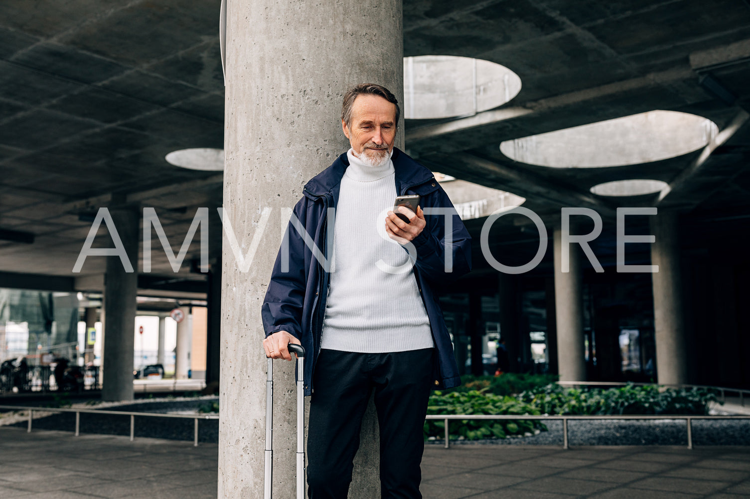 Senior man with rolling suitcase standing outdoors looking at mobile phone	