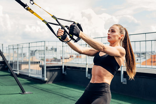 Young athlete doing push ups on a terrace. Woman exercising outdoors with suspension straps.