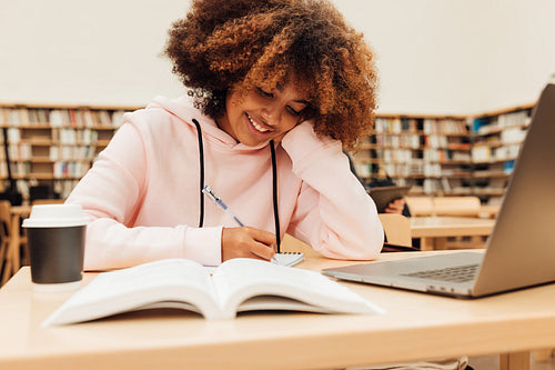 Smiling student writing in library. Girl with curly hair sitting at desk and learning.