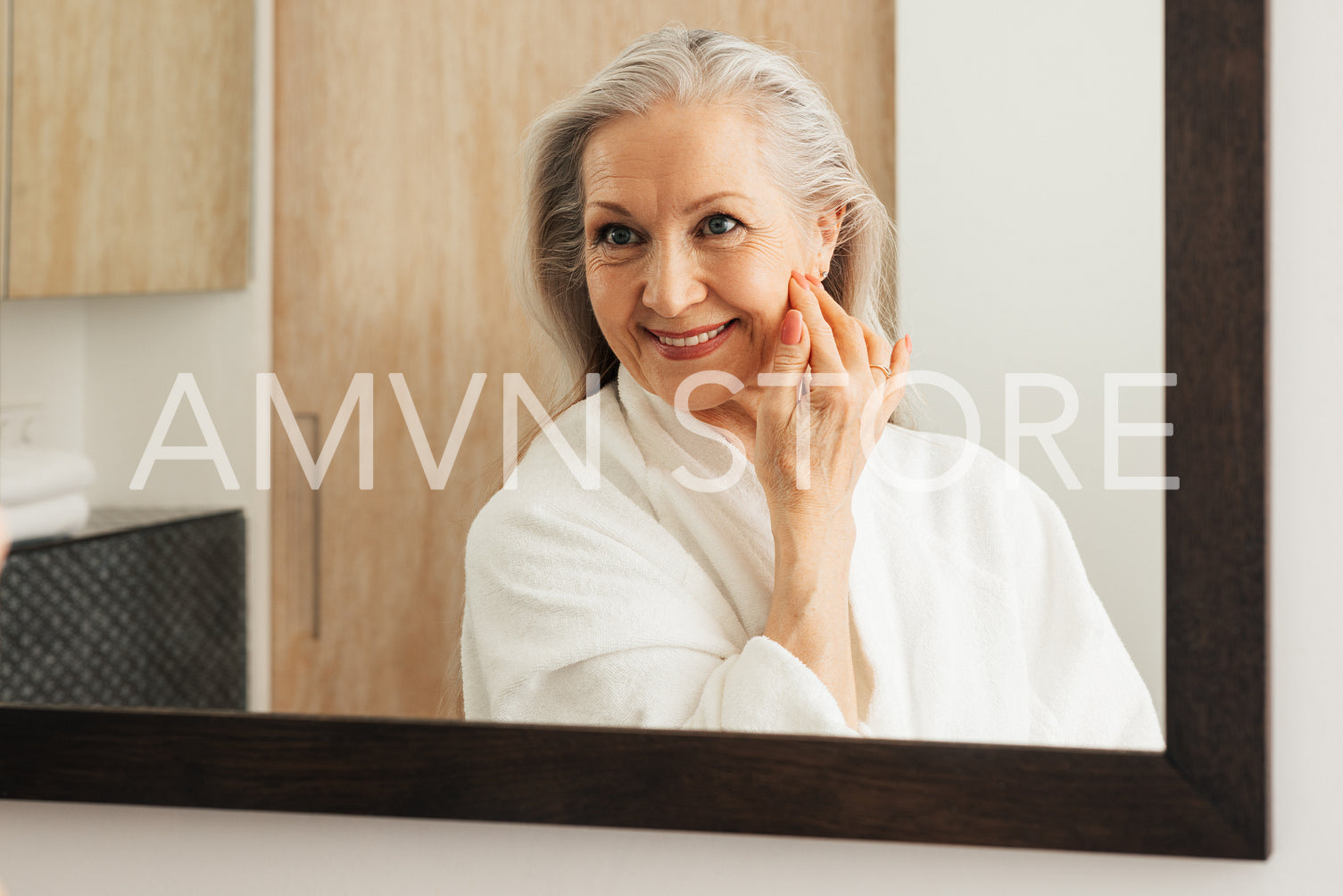 Mature woman with long grey hair looking at a mirror in a bathroom. Senior female in bathrobe adjusting her hair in front of a mirror.