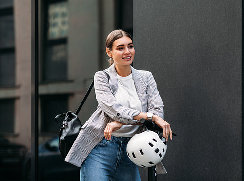 Businesswoman with backpack leaning on the handlebar of electric scooter looking away while standing at a black wall