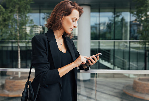 Side view of a woman in formal wear typing on a smartphone. Middle-aged female holding smartphone and standing outdoors.