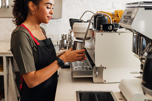 Side view of a female barista in apron standing at coffee machine and holding frothing pitcher