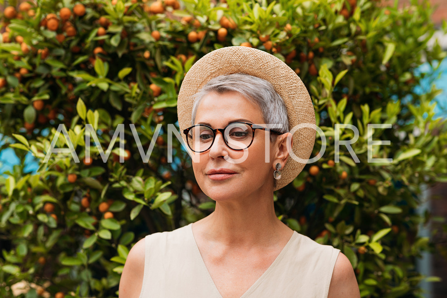 Stylish mature woman with grey hair wearing a straw hat and looking down while standing outdoors