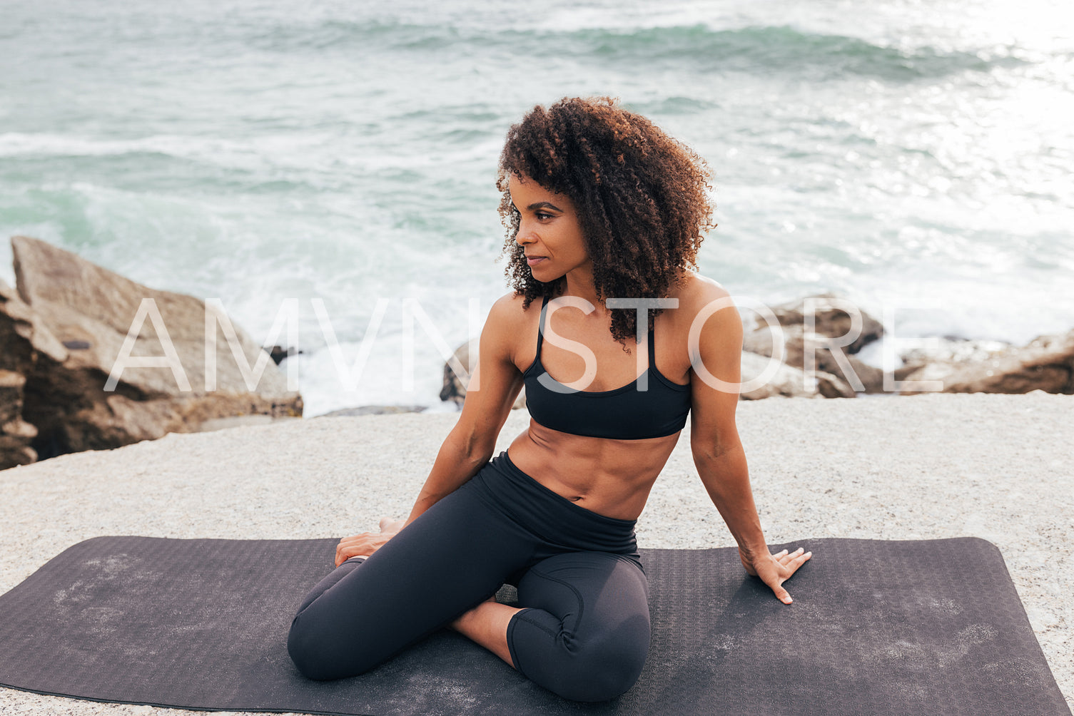 Muscular woman in sportswear sitting on mat by ocean. Female relaxing after workout at coast.