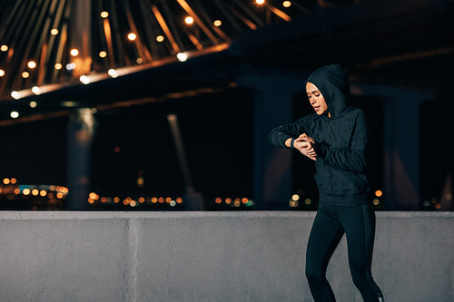 Young sportswoman looking at smartwatch while running at night outdoors