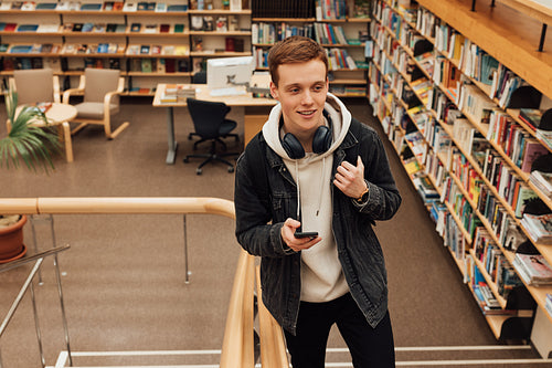 Smiling guy with smartphone in library. Student in casuals standing on stairs in university library.