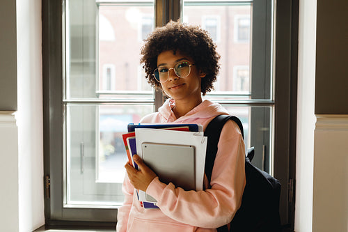 Beautiful young woman with books and digital tablet standing at window in campus