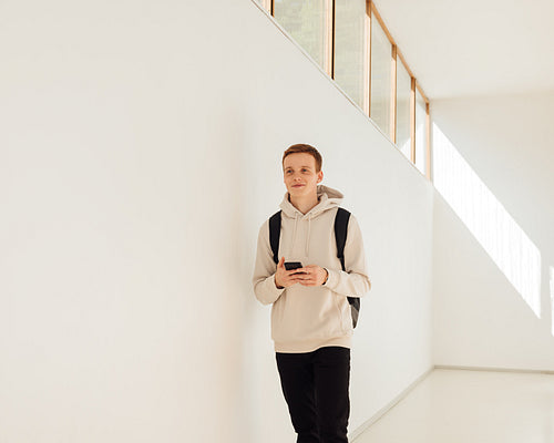 Cheerful student walking in corridor with smartphone. Young man in casuals walking in college building.