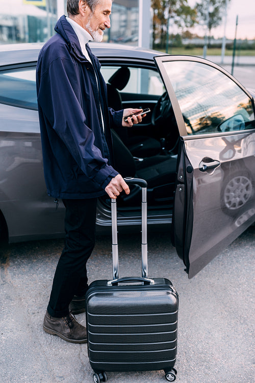 Side view of a mature tourist standing near the car and looking on cell phone