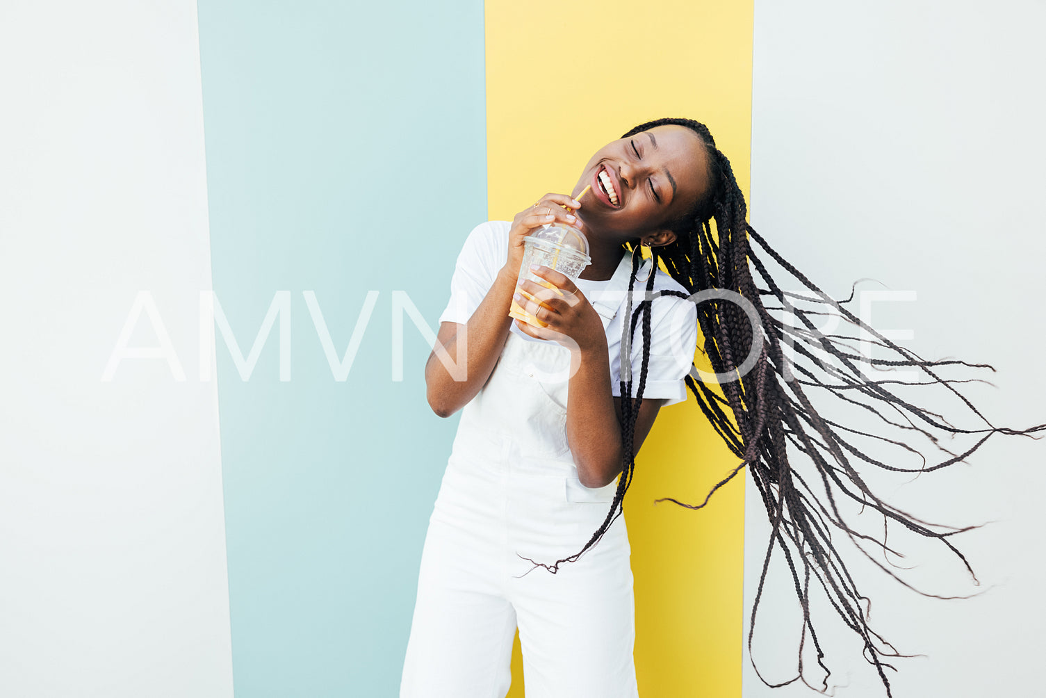 Cheerful woman with closed eyes drinking orange juice while dancing at a wall with blue and yellow stripes