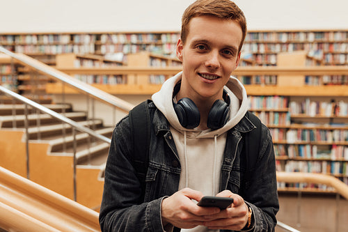 Portrait of a handsome student in casuals standing in library with smartphone