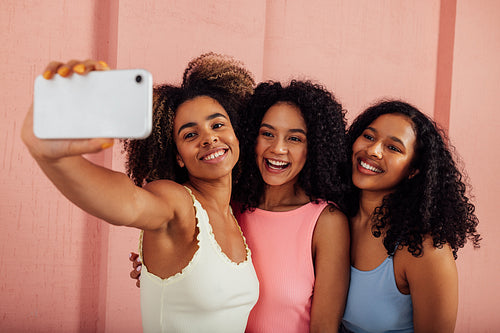 Young women in bright casuals standing together and taking selfie on a mobile phone. Happy females having fun against pink wall outdoors.