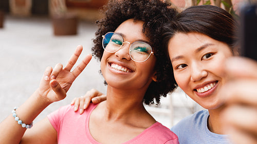 Two happy friends looking at smartphone and taking selfie, sitting in outdoor cafe