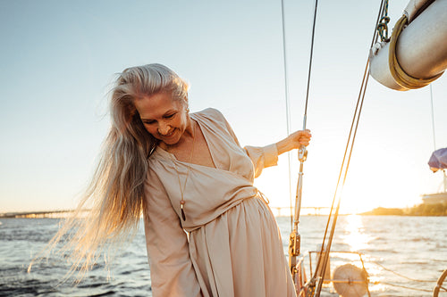 Beautiful mature woman with long gray hair standing on a sailboat and looking down at sunset